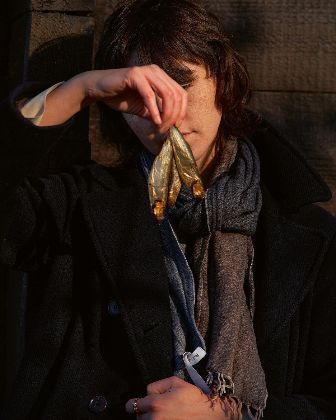 A person holding three dried fish near their face outdoors. The subject wears a dark coat and patterned scarf against a wooden background.