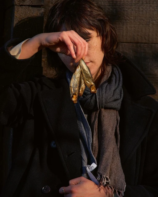 A person holding three dried fish near their face outdoors. The subject wears a dark coat and patterned scarf against a wooden background.