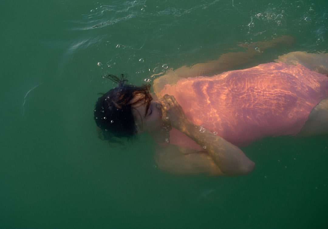 Underwater view of a woman with dark hair wearing a pink swimsuit, seen from above in a turquoise-tinted body of water with ripples.