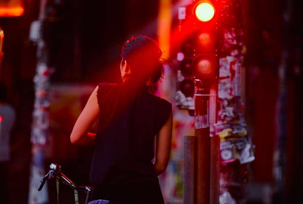 A woman on a bicycle waits at a red light at dusk. The scene is illuminated by the bright red stoplight, creating a dramatic effect.