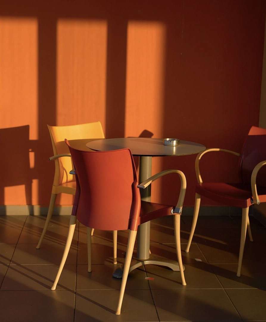 Sunlit seating area featuring a round table surrounded by three chairs with a solid orange wall, creating a warm, inviting atmosphere.