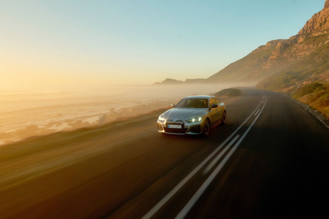 Silver BMW driving on coastal road at golden hour with fog and mountains, aerial perspective. Automotive beauty shot of the electric car.