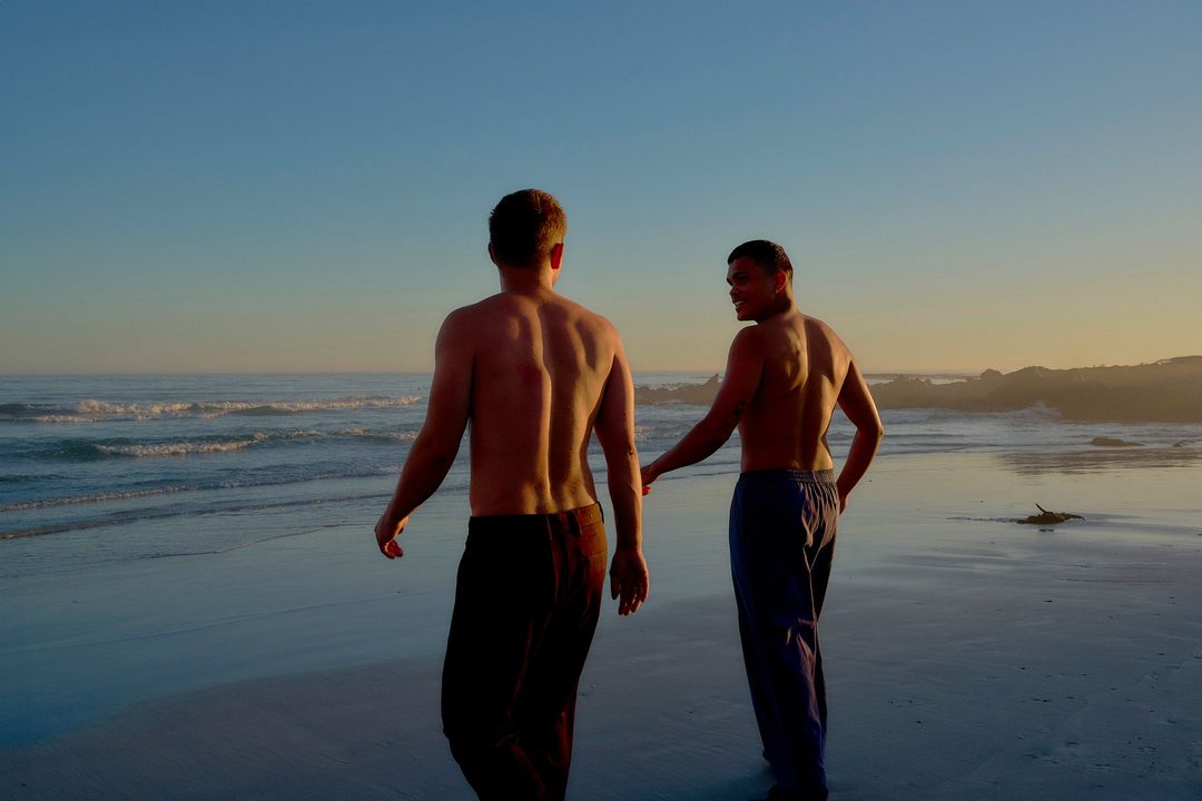 Two shirtless adult men walking hand-in-hand on a sandy beach at sunset, waves gently crashing in the background under a clear sky.