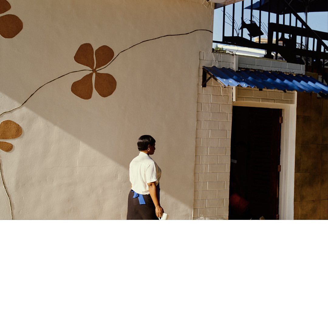 Adult woman stands near wall decorated with floral motif, light washing over architectural detail, creating a bright, sunny outdoor scene.