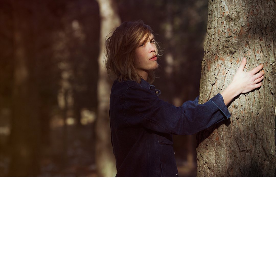 Woman with short hair embraces a tree in a forest setting, wearing a denim jacket, bathed in soft light, and surrounded by blurred trees.