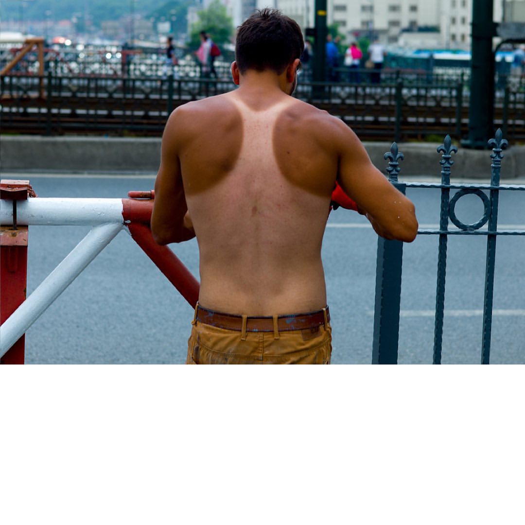 Adult male with visible tan lines standing on a city street behind metal railing in daylight with a blurred background of buildings.