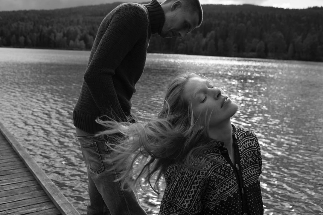 Monochrome shot of a blond woman with her head tilted back and eyes closed, standing on a wooden pier with a man near a lake.