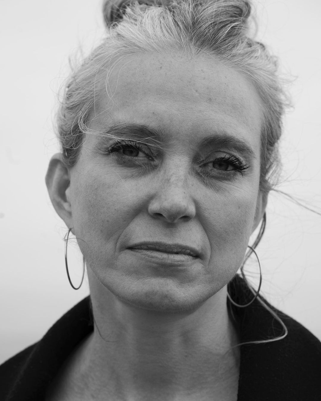 Black and white portrait of an adult woman with grey hair in a high bun, wearing hoop earrings and a dark coat, against a light background.