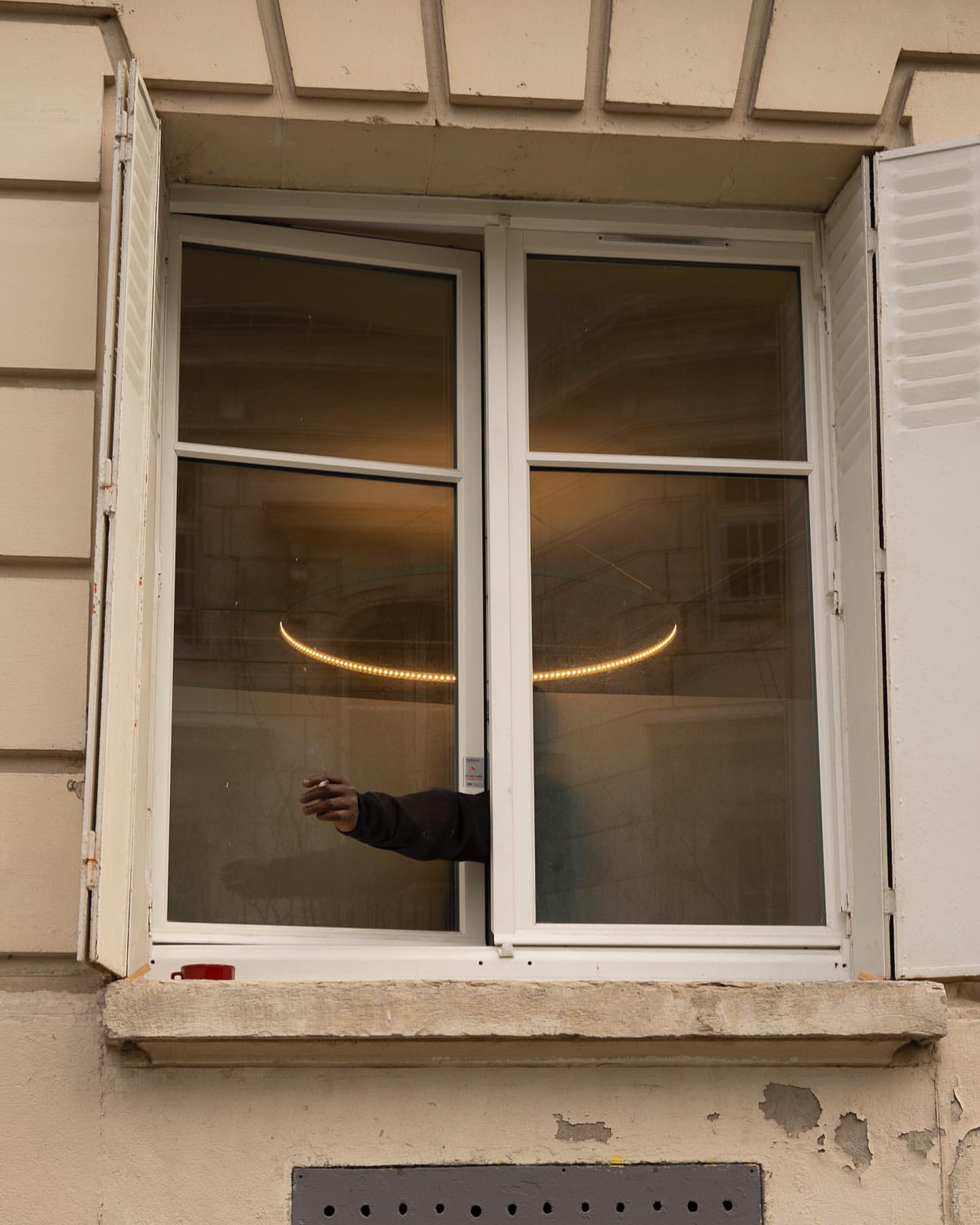 Exterior building shot focusing on a window with open shutters and an arm reaching out holding a cigarette. A light is visible inside.