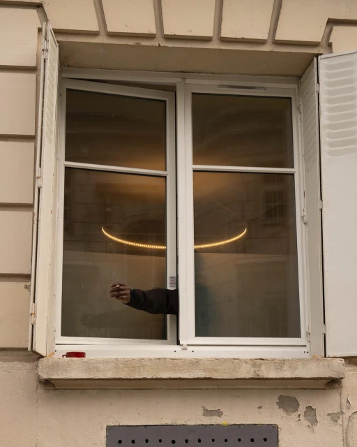 Exterior building shot focusing on a window with open shutters and an arm reaching out holding a cigarette. A light is visible inside.