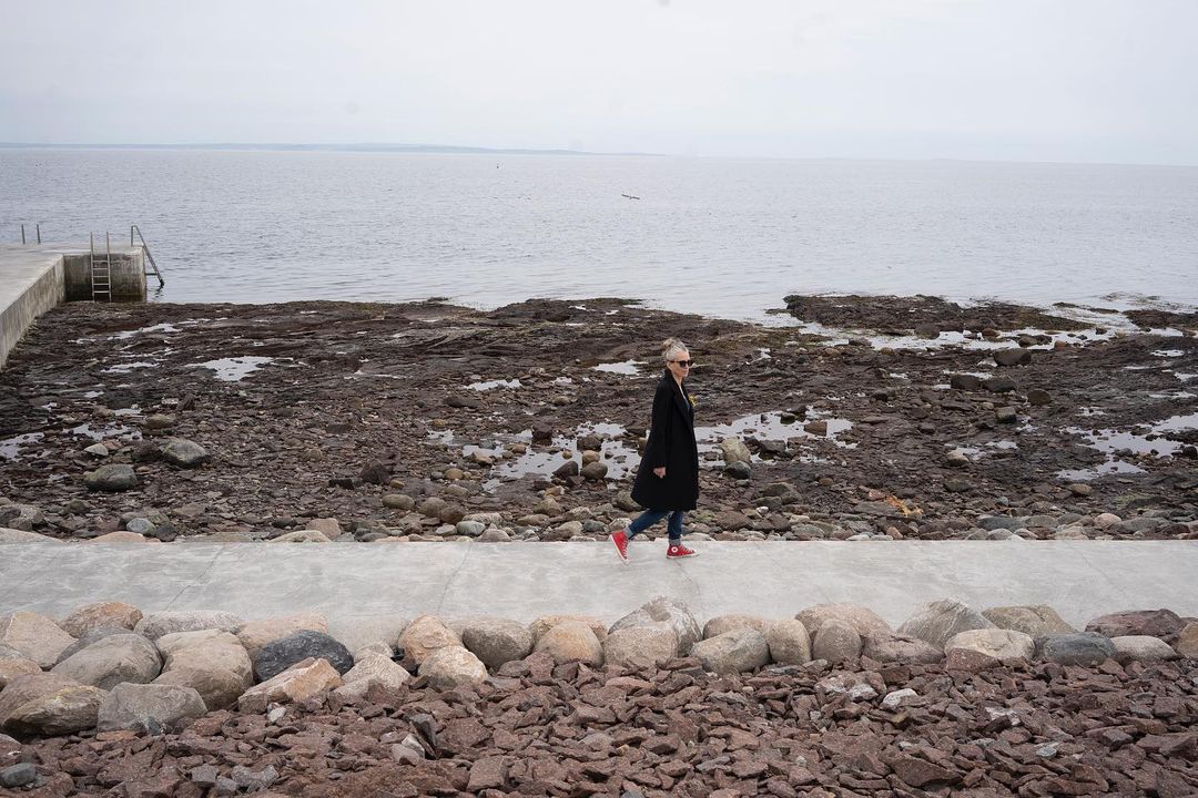 Adult woman walking along a concrete path next to a rocky beach and ocean under an overcast sky, wearing a black coat and red shoes.
