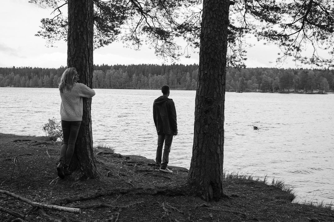 Black and white shot of two people standing near a lake, surrounded by trees, with one person hugging a tree trunk.