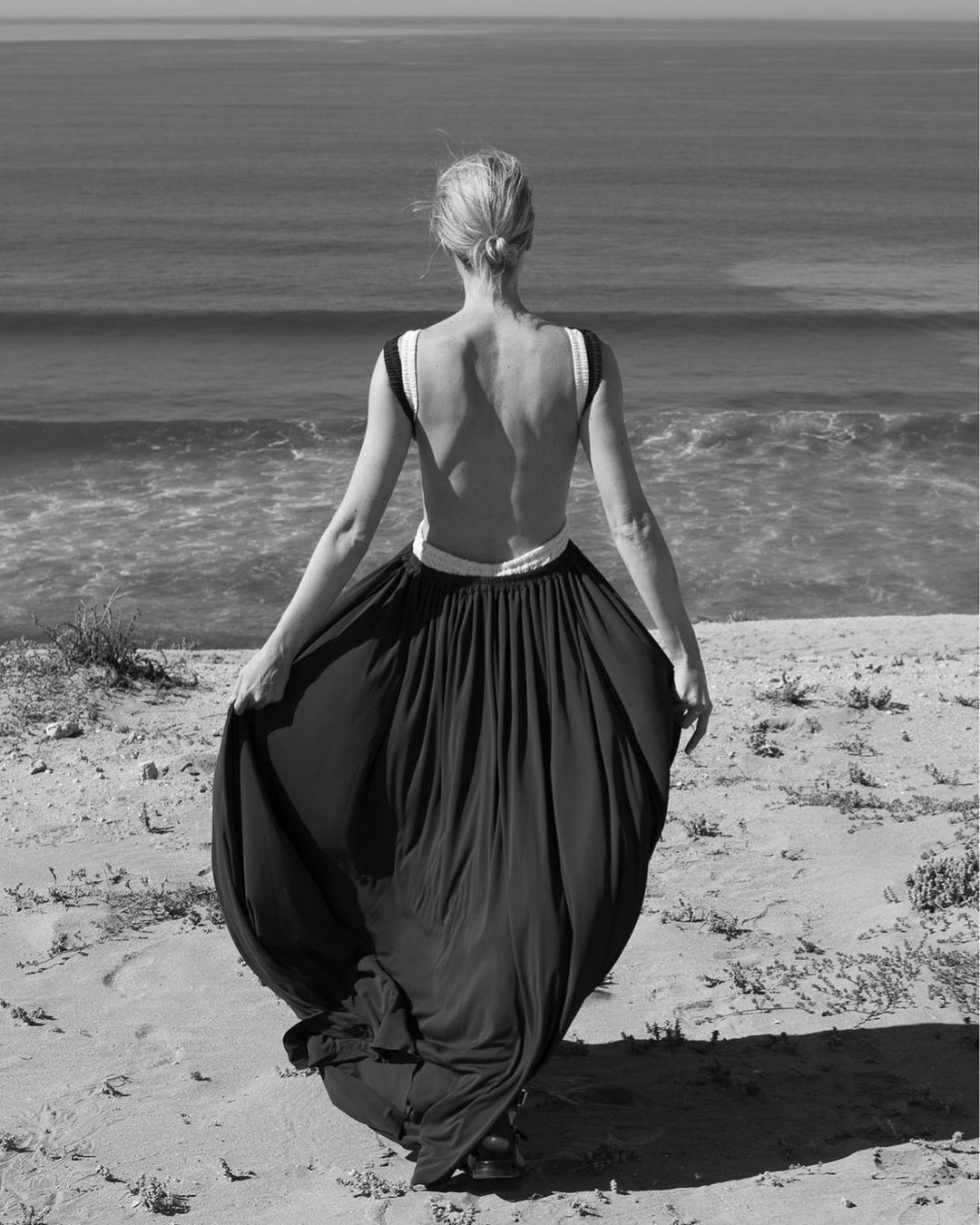 Black and white image of a woman in a long black dress standing on a sandy hill looking out at the ocean waves.