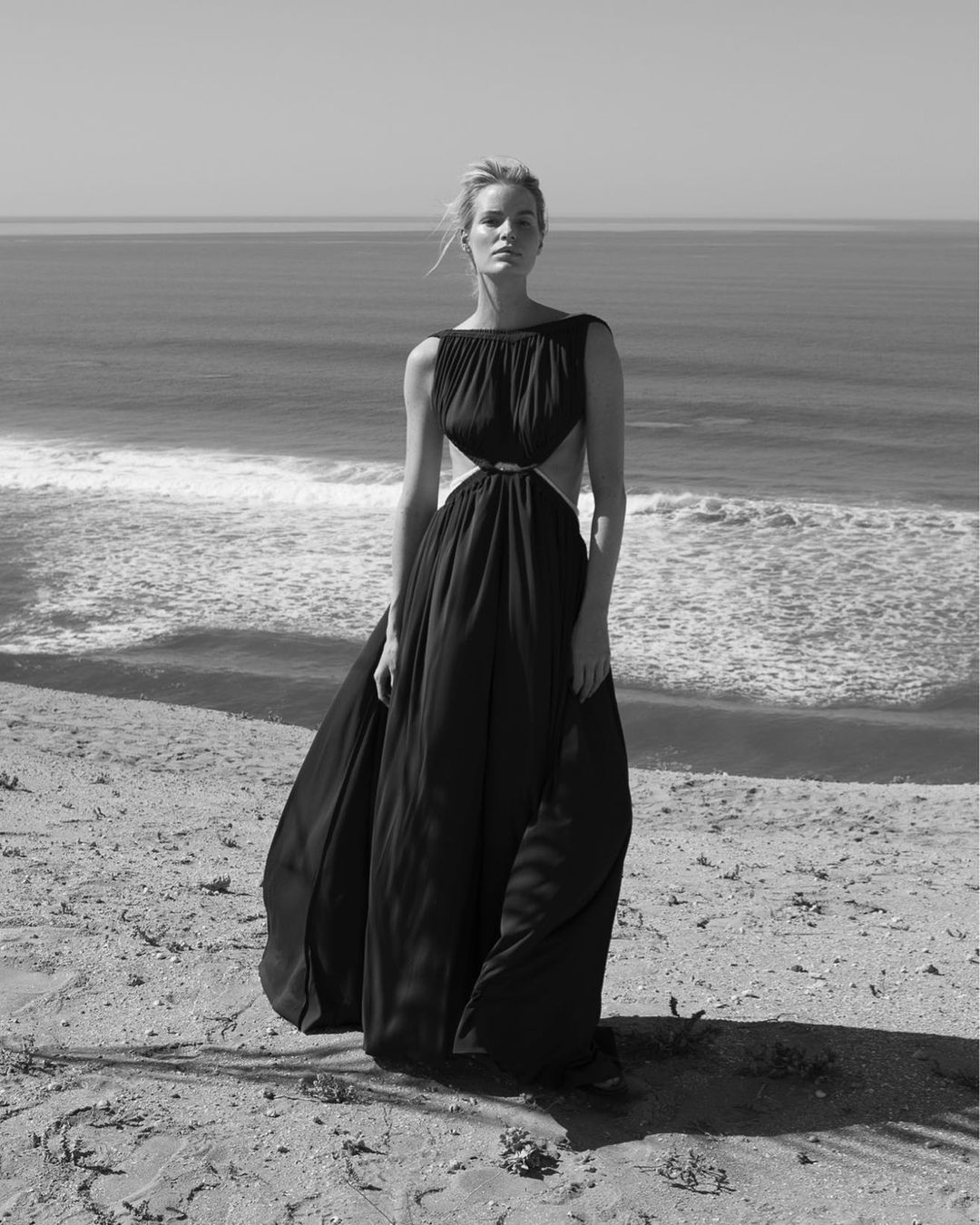 Woman stands in a black flowing gown on a sandy shore with an ocean backdrop, under a bright sky, creating a minimalist monochrome aesthetic.