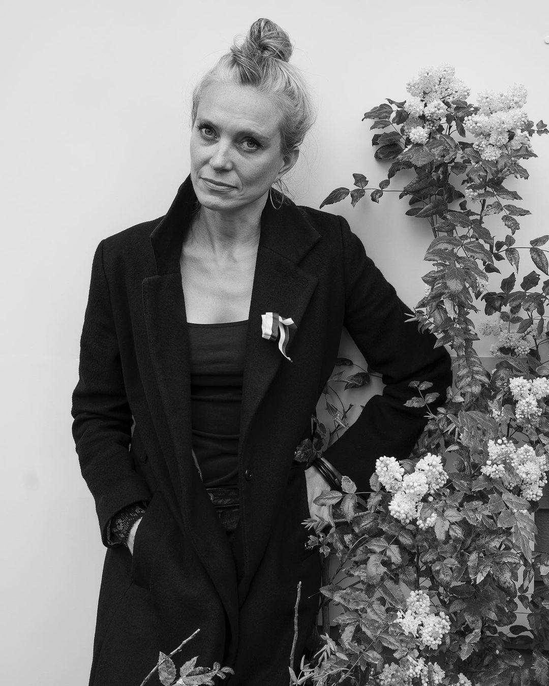 Black and white portrait of an adult woman with a topknot hairstyle, wearing a dark coat, standing near a flowering vine.