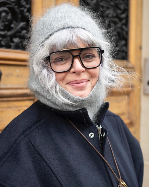 Portrait of an adult woman with gray hair and glasses, wearing a wool hat and navy coat, standing outside against a neutral background.