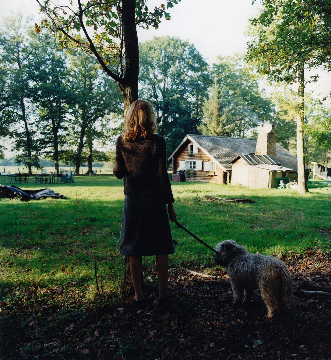 A woman and a dog in a grassy, wooded area in front of a rustic house. The scene is bathed in soft, natural light.