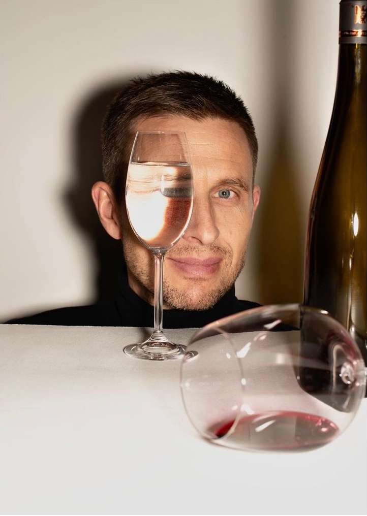 Man looking through a wine glass held in front of his eye, with a wine bottle and a spilled glass on a table.