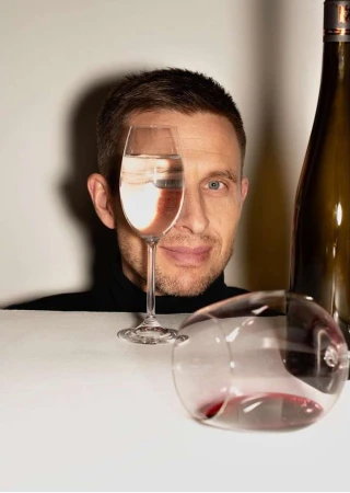 Man looking through a wine glass held in front of his eye, with a wine bottle and a spilled glass on a table.