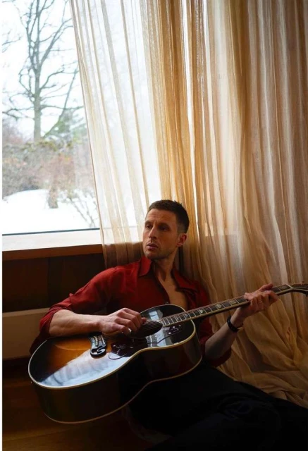 Man playing acoustic guitar indoors next to a window with beige curtains, wearing a red open-collar shirt and black pants in a relaxed pose.