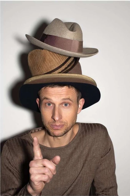 Man wearing a stack of three hats in a studio setting. He points his index finger up while wearing a brown long-sleeved shirt.