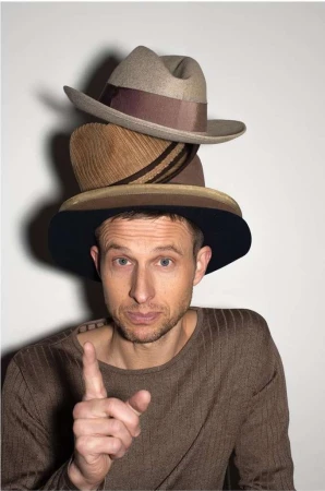 Man wearing a stack of three hats in a studio setting. He points his index finger up while wearing a brown long-sleeved shirt.