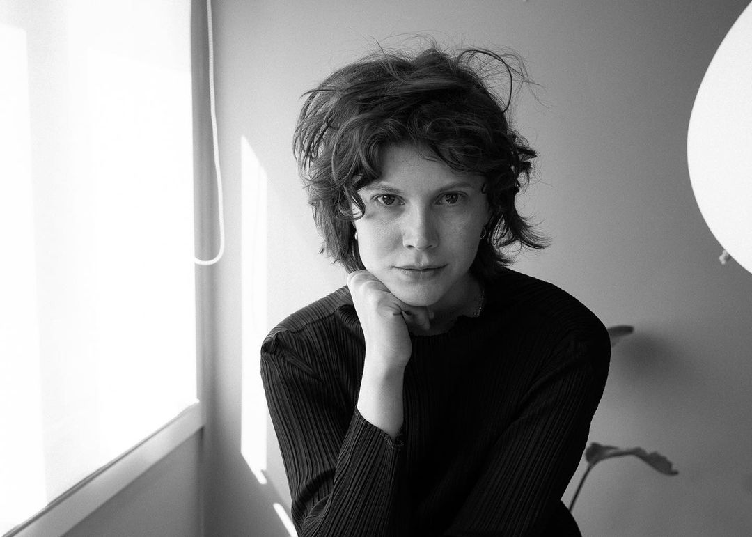 Monochromatic portrait of a woman with short curly hair in a dark pleated top, softly lit by a window, in an indoor setting.