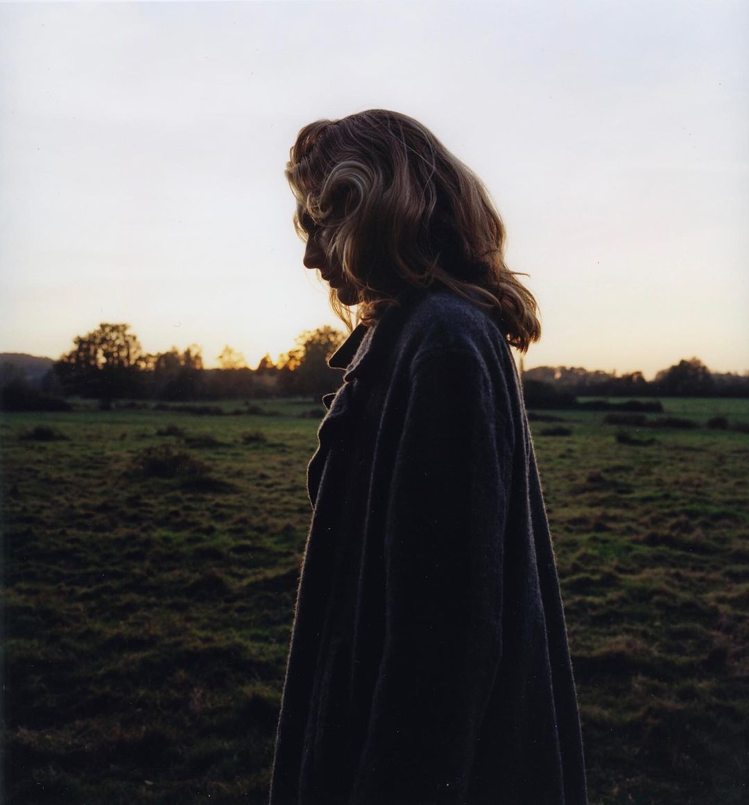 Woman in coat stands in grassy field against sunset sky. The woman has wavy hair and is gazing downward in this outdoor shot.