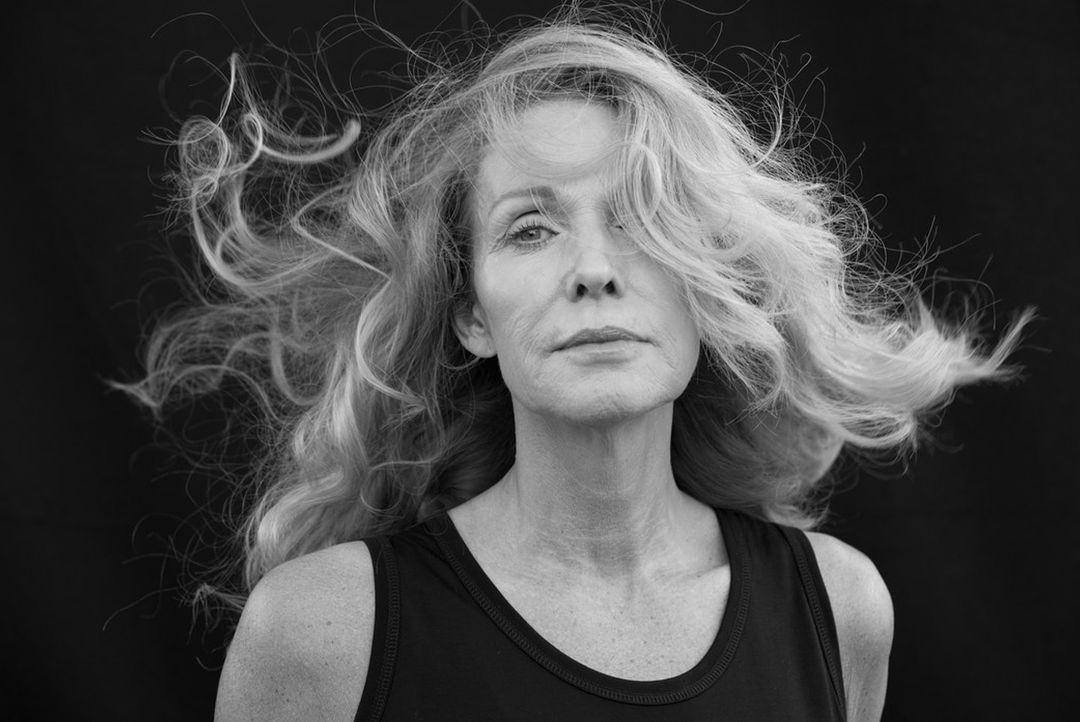 Black and white portrait of an older woman with blonde wavy hair wearing a tank top against a black background in a photo studio.