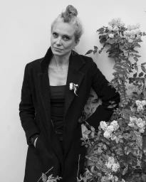 Portrait of an adult woman in a dark coat standing next to a flowering plant, photographed by Lars Botten