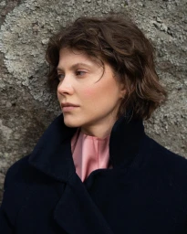 Portrait of a woman with short dark hair wearing a dark coat and pink collar against a textured stone wall