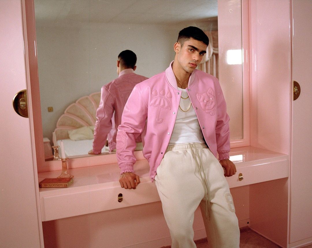 A young adult male in a pink jacket poses in a pink room, a mirror reflecting a bed, creating a cohesive and fashionable portrait.