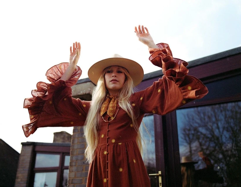 A blonde woman in a rust-colored dress with star patterns raises her arms in front of a window, wearing a hat and scarf.