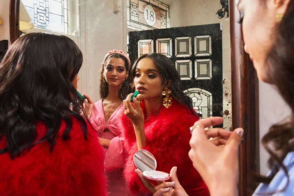Two women apply makeup in front of a mirror. One woman in red applies lipstick and holds a compact mirror. Interior setting.