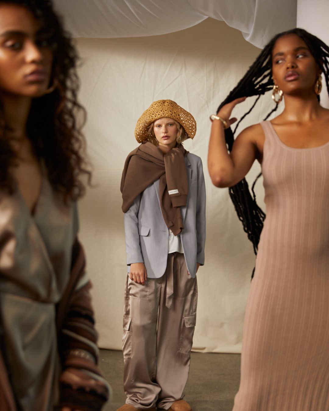 Three women model neutral-toned clothing in a studio setting, including a dress, blazer, scarf, and cargo pants, under soft lighting.