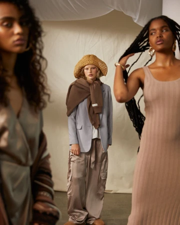 Three women model neutral-toned clothing in a studio setting, including a dress, blazer, scarf, and cargo pants, under soft lighting.