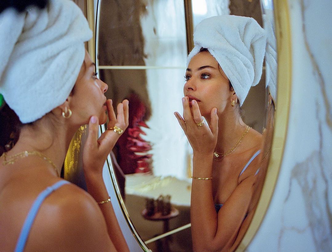 Woman with a towel on her head is applying skincare in front of a bathroom mirror, accessorized with gold jewelry and a blue top.