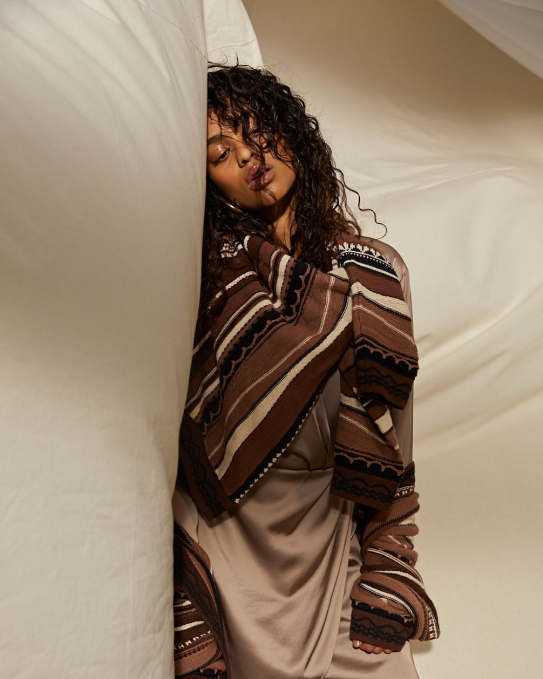 Woman with curly hair wearing a brown and beige patterned wrap and dress, posing against a draped white fabric background.