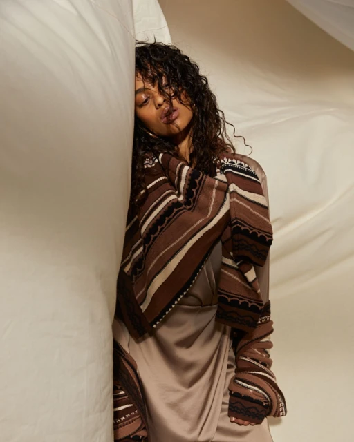 Woman with curly hair wearing a brown and beige patterned wrap and dress, posing against a draped white fabric background.