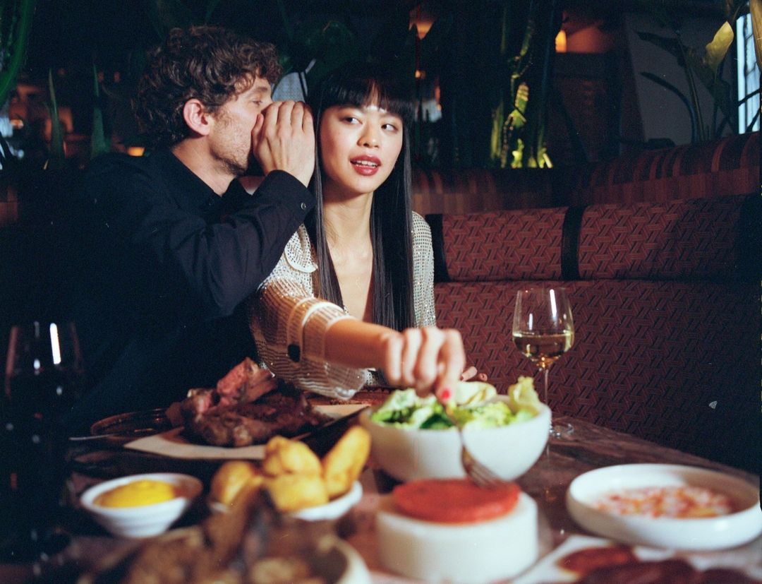An Asian woman and a Caucasian man at a restaurant, with the man whispering in the woman's ear, surrounded by food and drinks.