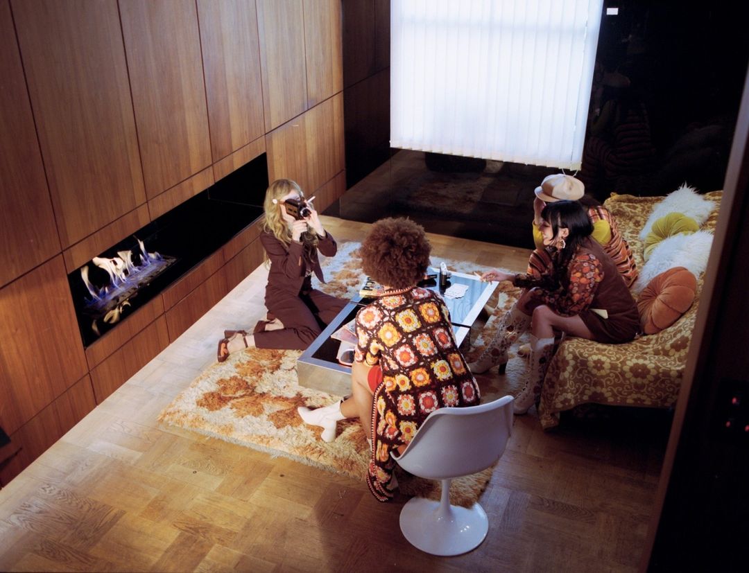 Four people in retro clothing gather in a 70s-style room featuring wood paneling, a fireplace, shag rug, and patterned sofa, indoor.