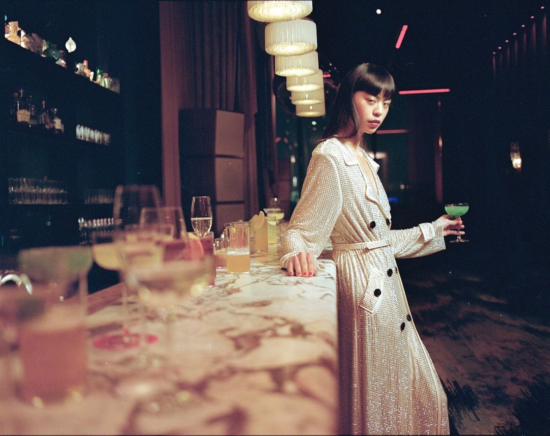 Elegant woman in a shimmering trench coat at a bar, holding a green cocktail; marble countertop, and soft lighting create a sophisticated scene.