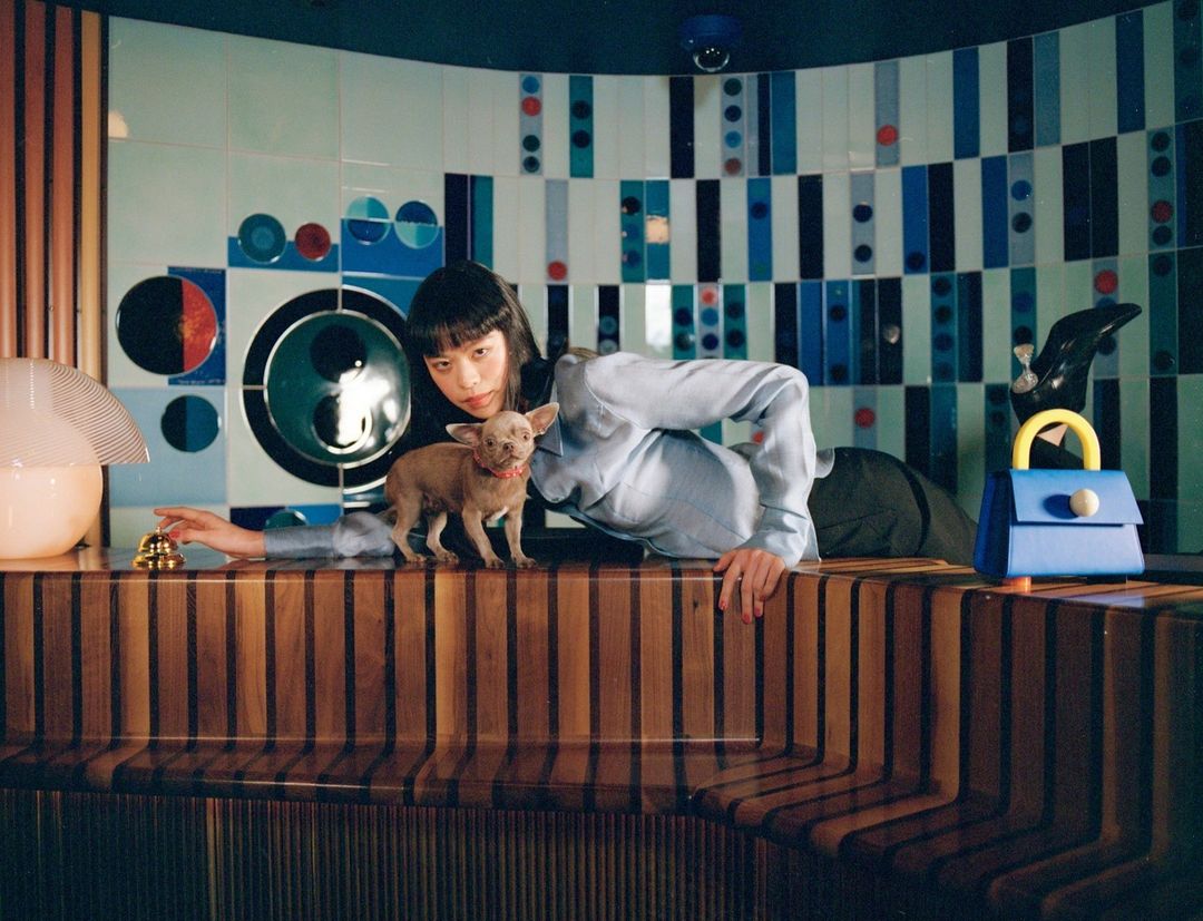 Woman poses with a small dog behind a textured counter, with purse and architectural tiles in the background, indoor fashion photography.