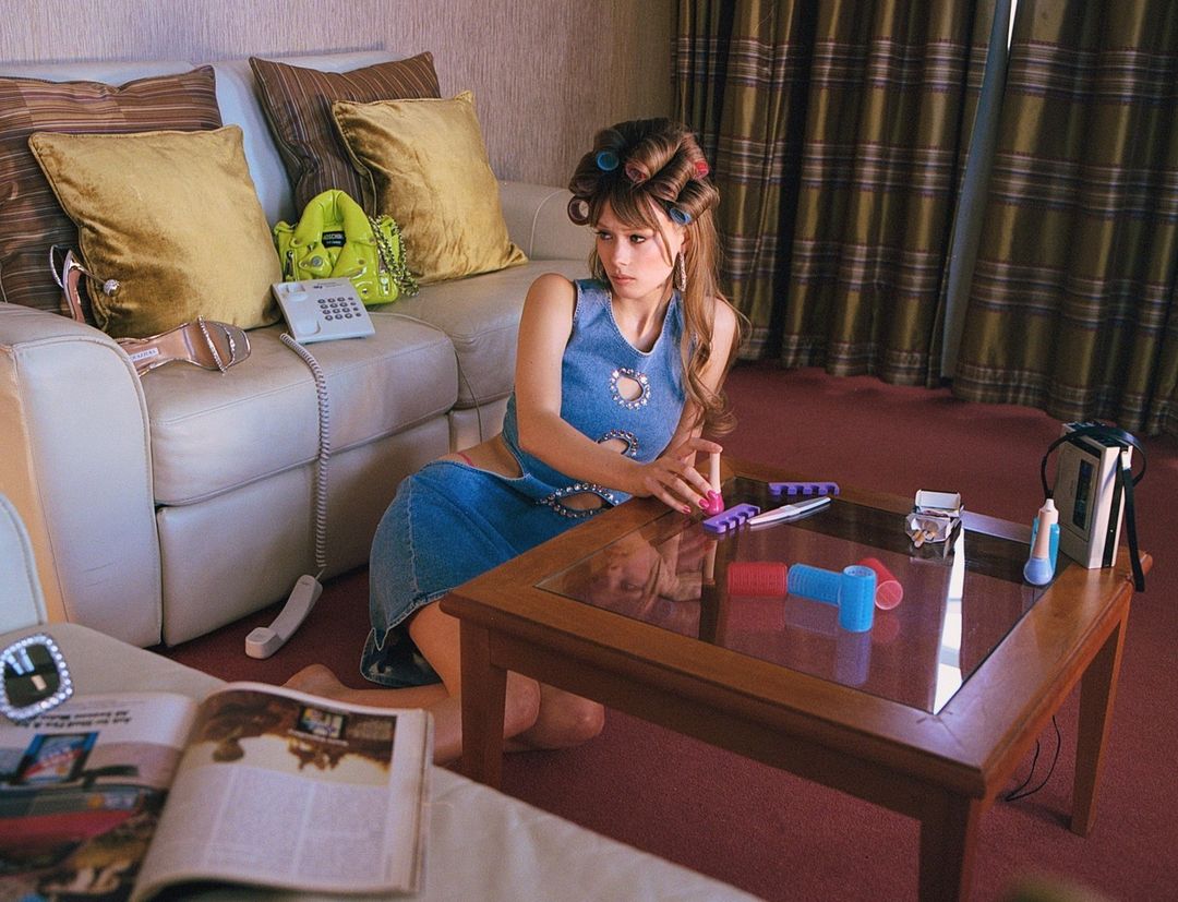 Young woman sits on the floor in a blue jean dress and hair rollers, next to a coffee table with a magazine and beauty products.