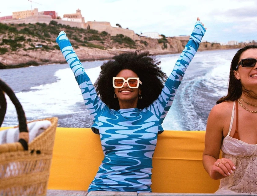 Two women on a boat, with one woman in a blue patterned dress raising her arms, smiling with sunglasses. Coastal architecture in background.
