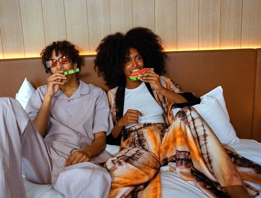 Two stylish individuals are sitting on a bed, smiling and eating watermelon, showcasing fashion-forward eyewear and clothing in an indoor setting.