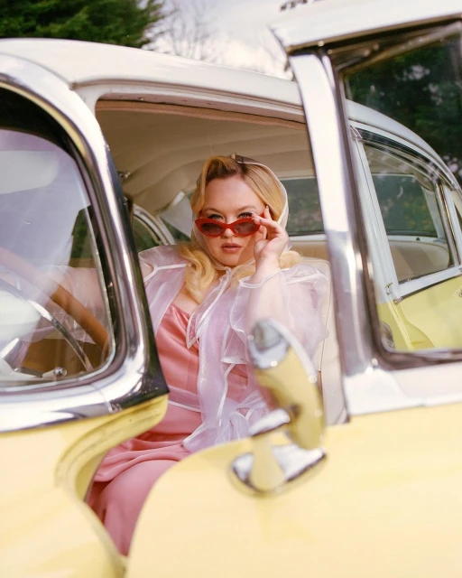 Blonde woman in retro sunglasses sits in a vintage yellow car with its door open, wearing a pink dress and sheer white shawl.