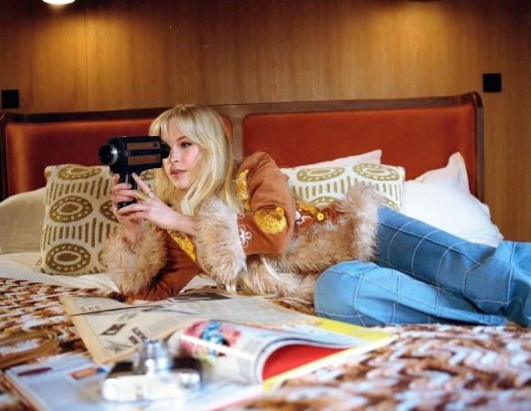 Blonde woman holding a vintage movie camera while lying on a bed with magazines, photographed by Laura Allard-Fleischl