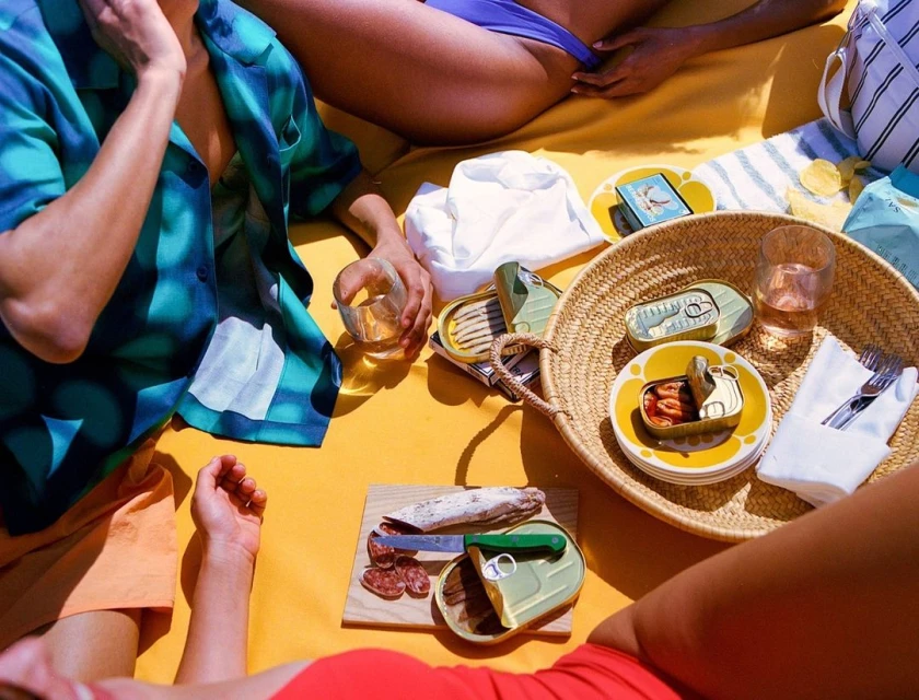 Overhead shot of people relaxing on a yellow surface with canned seafood, cheese, sausage, and drinks for a picnic or snack.