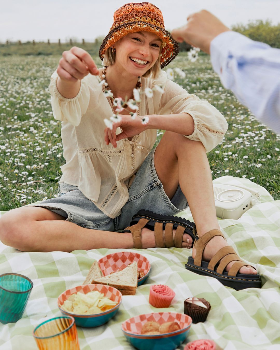 Young woman in a crochet hat smiles, sitting on a picnic blanket in a daisy field, throwing flower petals, with a picnic spread.